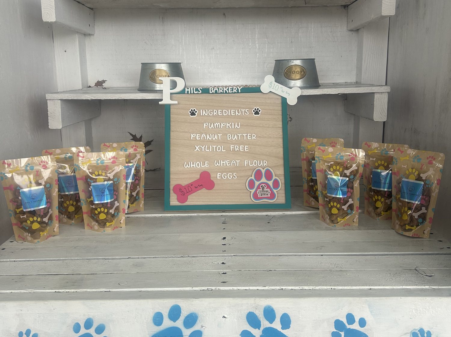 The Phil's Barkery farmstand: bags of treats either side of a board listing the ingredients, with dog bowls on the shelf above and blue paw prints painted along the counter.