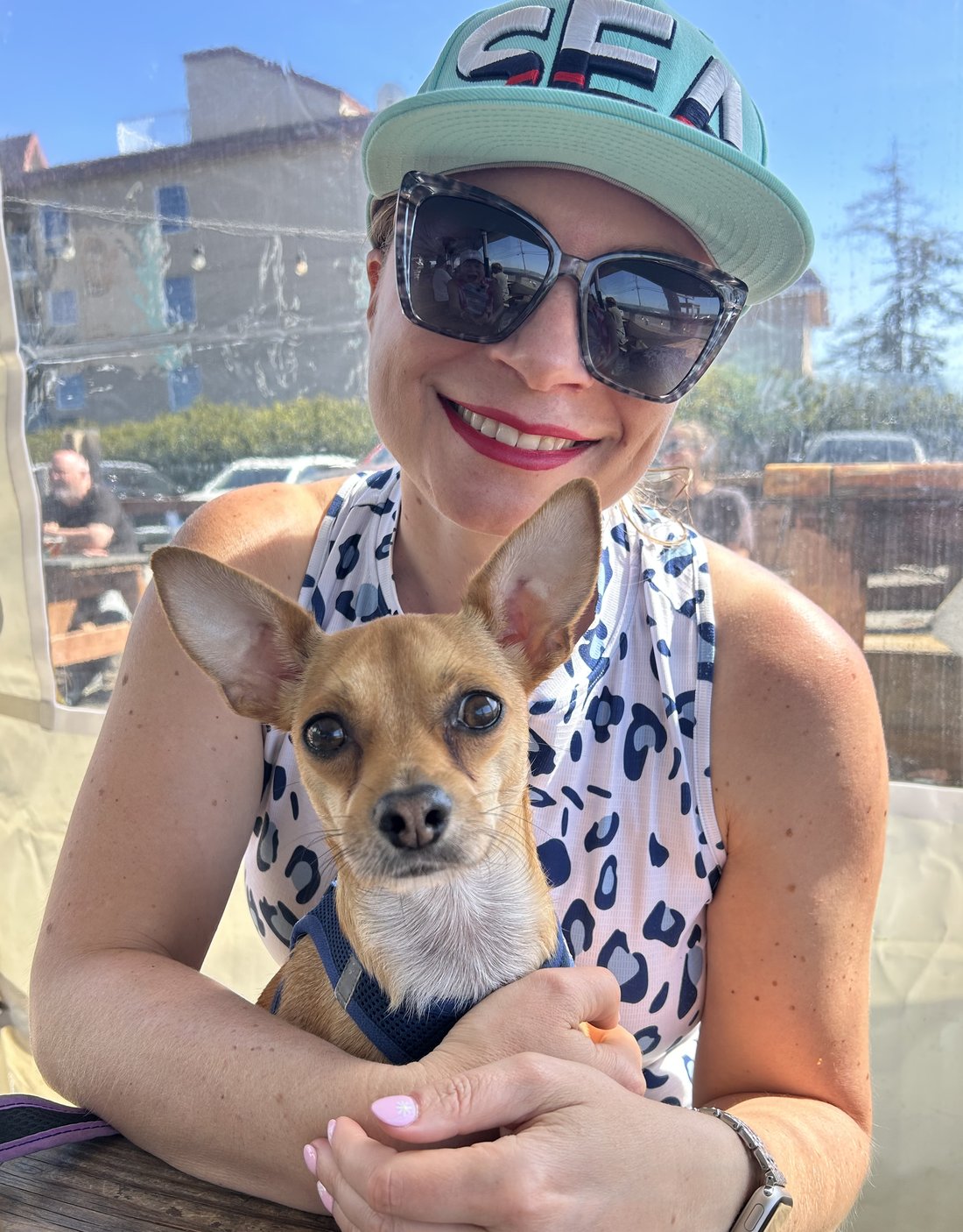 Phil, a tan chihuahua mix in a navy harness, being held at an outdoor table on a sunny day by the person who bakes the treats. Both are looking at the camera.